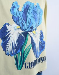 Κοντινό πλάνο σε μπεζ ύφασμα με μεγάλο λουλούδι ίριδας σε μπλε και λευκό χρώμα και το κείμενο "GIARDINO MICHELANGELO BUONARROTI, FIRENZE FI, ΙΤΑΛΙΑ" από κάτω.