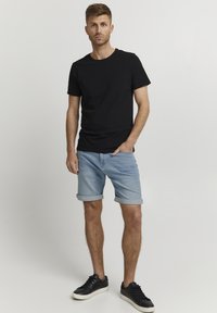 Svart t-shirt med rund halsringning, ljusblå denimshorts med uppvikta ben och svarta sneakers. Mannen står med en hand i fickan. Bakgrunden är enfärgad.