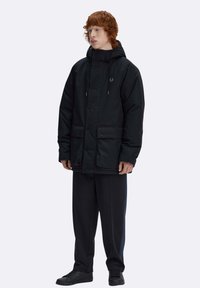 Giacca imbottita nera con cappuccio, dotata di bottoni a pressione, due tasche frontali e un logo sul petto, abbinata a pantaloni e scarpe nere.