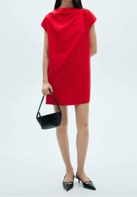 Rotes Shiftkleid mit drapiertem Ausschnitt, kurzen Ärmeln und lockerem Schnitt. Kombiniert mit einer schwarzen Handtasche und schwarzen spitzen Pumps.