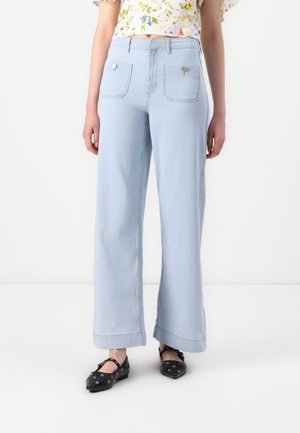 Jeans larges bleu clair à taille haute, avec deux poches arrière et des accents métalliques, ainsi qu'un ourlet propre. Associés à un haut floral et des ballerines noires.