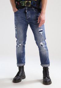 Modré džíny s opotřebováním a dírami, tapered střih, s ohrnutými nohavicemi, v kombinaci s černými šněrovacími botami. Měkká textura s bledou úpravou.