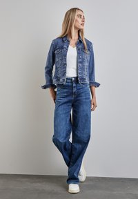 Blå denimjacka med lätt slitage, bärs över en vit V-ringad topp. Matchas med vida blå jeans och vita sneakers. Enkelt design.