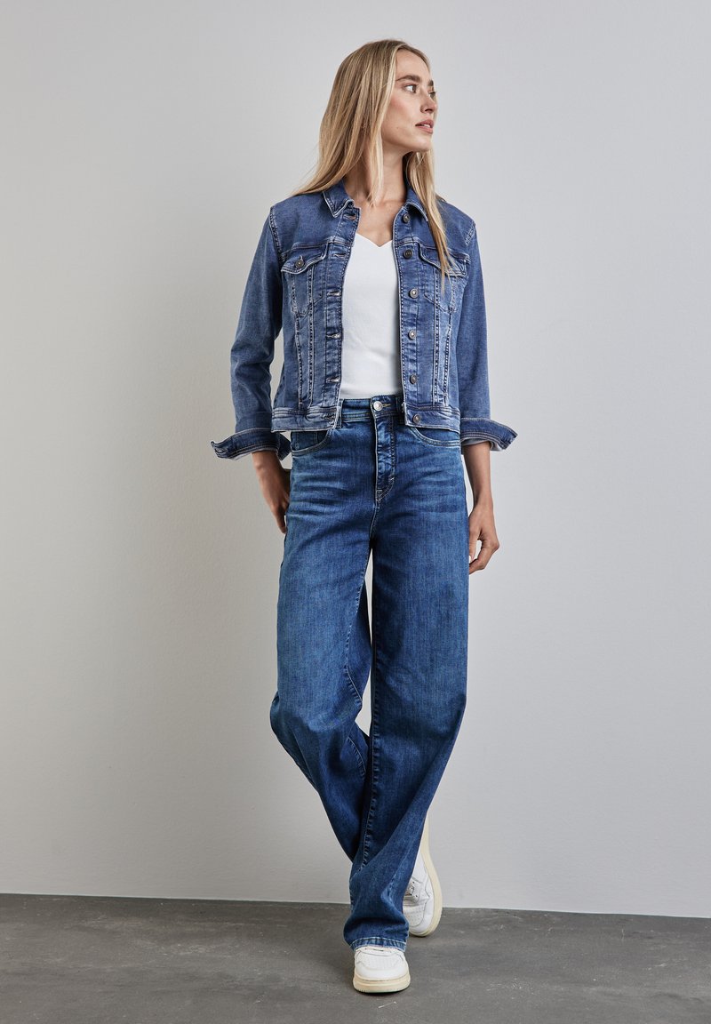 Blå denimjacka med lätt slitage, bärs över en vit V-ringad topp. Matchas med vida blå jeans och vita sneakers. Enkelt design.