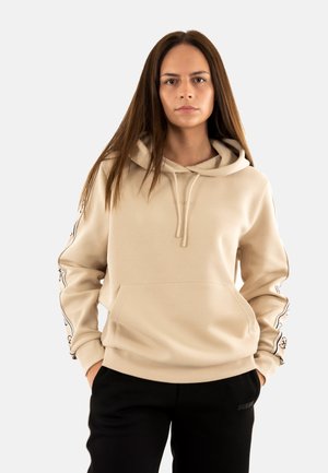 Sudadera beige con un bolsillo frontal, cuello con cordón y acentos estampados en las mangas. Hecha de una tela suave y texturizada. Combinada con pantalones negros.