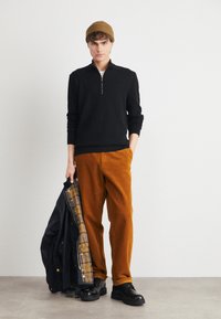 Black zippered sweater, orange corduroy pants, and black shoes. Holds a black jacket with a plaid lining. Brown knit beanie. Neutral backdrop.