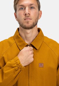 Hořčicově žlutá bunda s vysokým límcem, zapínáním na zip a elastickými manžetami. Na přední straně se nachází malý hnědý logový štítek.