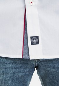 Weißes Hemd mit Knopfleiste und roten Akzenten sowie blauen Denim-Details, mit einem Logo-Patch am Saum. Kombiniert mit blauen Jeans.