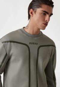 Szürke kapucnis pulóver zöld részletekkel és "GUESS" logóval. Kerek nyakkivágással rendelkezik, sima textúrával és minimalista dizájnvonalakkal.