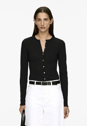 Black long-sleeve top featuring a round neckline, front buttons, and a fitted design, worn with white pants and a black belt.
