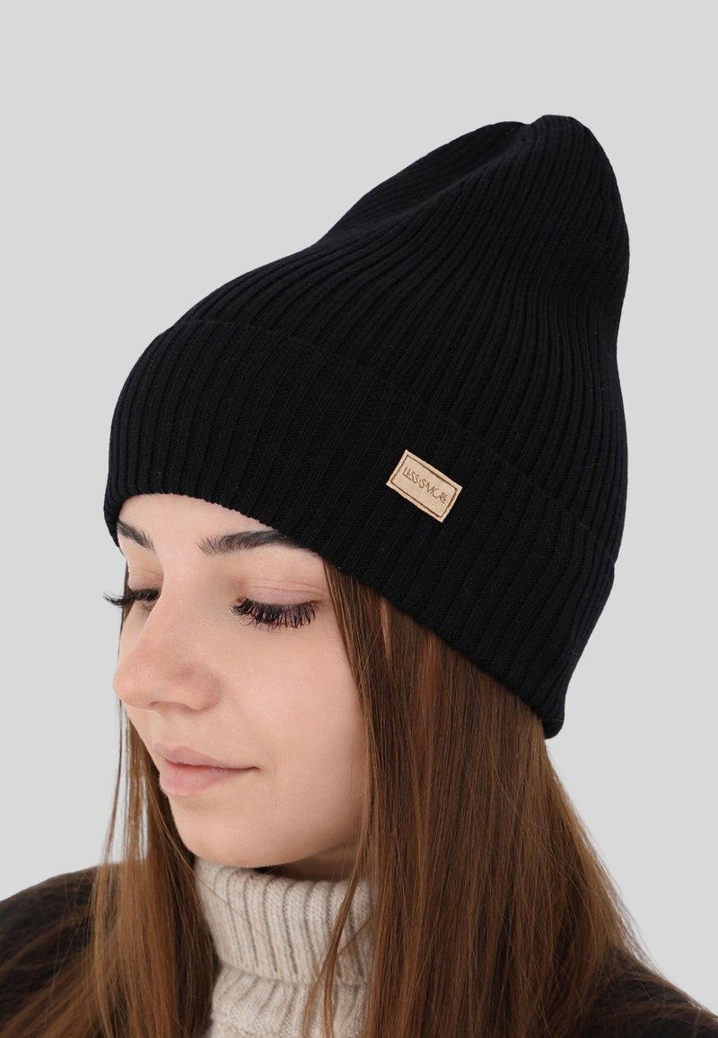 Jonge vrouw met lang bruin haar, die een geribbelde zwarte beanie en een beige coltrui draagt, licht naar beneden en naar links kijkend.