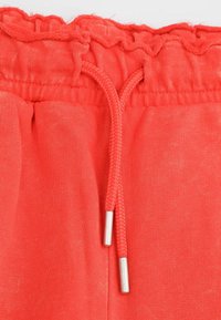 Elastyczny pas z falbaną na górnej krawędzi, regulowany sznurek w plecionym czerwonym materiale z metalowymi końcówkami w kolorze srebrnym. Tkanina jest miękka i lekka.