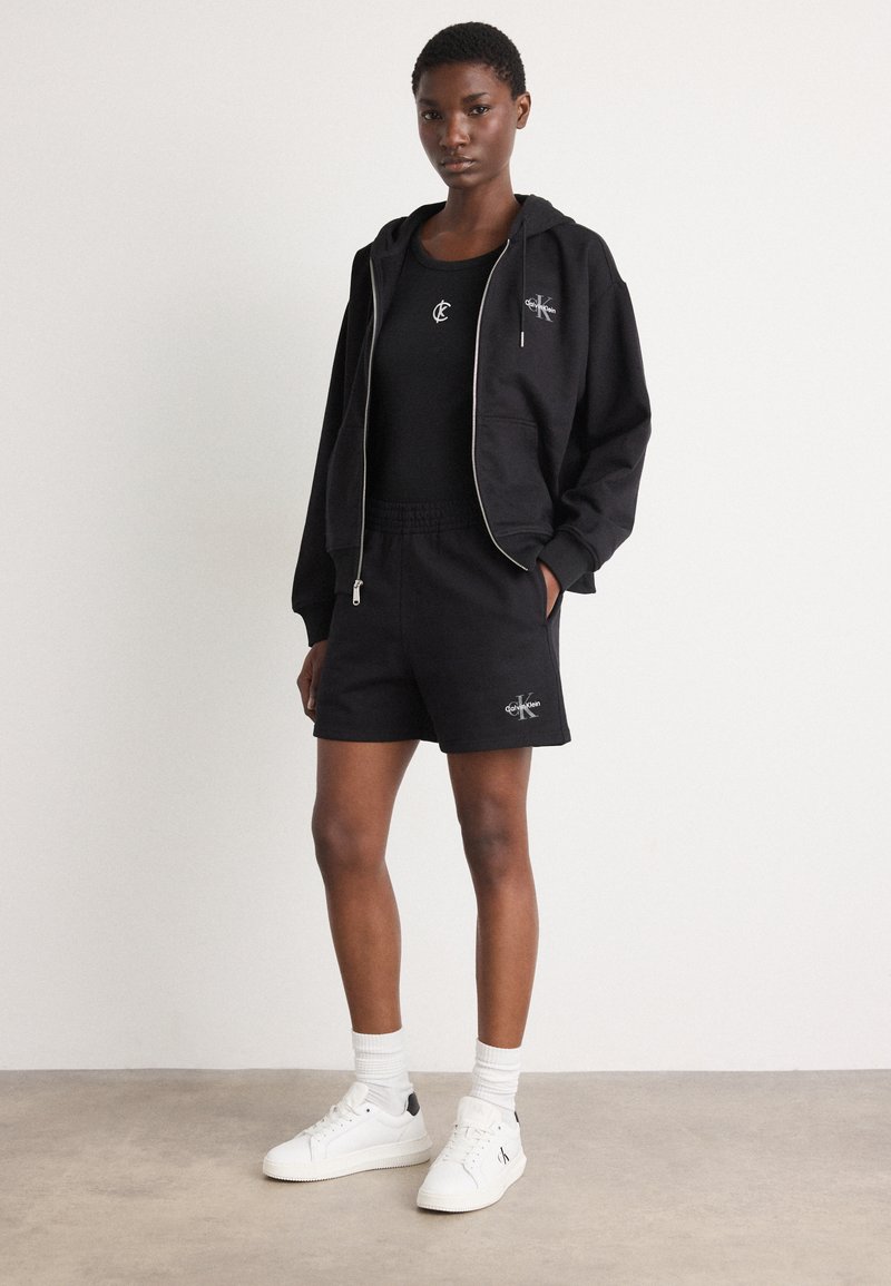 Zwarte katoenen hoodie met rits, zwart tanktop en zwarte shorts met een geborduurd logo. Witte sneakers met zwarte accenten en witte sokken.