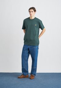 Tmavě zelené bavlněné tričko s krátkými rukávy a malou nášivkou, kombinované s volnými modrými džíny a hnědými koženými botami.