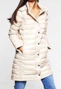Beige péřová bunda s prošívaným designem, která má límec, zapínání na knoflíky a boční kapsy. Vyrobena z lesklého izolačního materiálu.