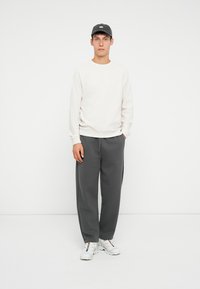 Sudadera de color crema claro, pantalones de chándal grises y zapatillas blancas. La sudadera tiene puños y dobladillo acanalados; los pantalones son de corte holgado. Lleva una gorra negra.