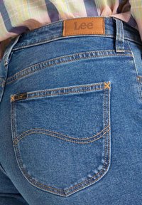 Pantalones vaqueros de mezclilla azul con costuras naranjas, que presentan un parche de cuero de la marca Lee y un diseño único en el bolsillo trasero.