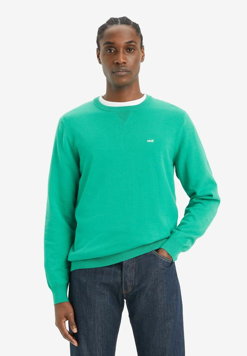 Suéter de cuello redondo en color turquesa con puños y dobladillo acanalados. Cuenta con un pequeño logo blanco en el pecho. Combinado con unos jeans de mezclilla oscura.