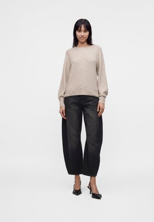 Jeune femme debout, portant un pull beige, un jean noir large et court, et des chaussures à talons hauts pointues noires sur un fond uni.