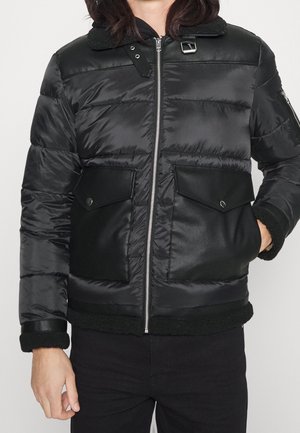 Person wearing black padded jacket with zip front, two large flap pockets, buckle collar, and one hand inside pocket.