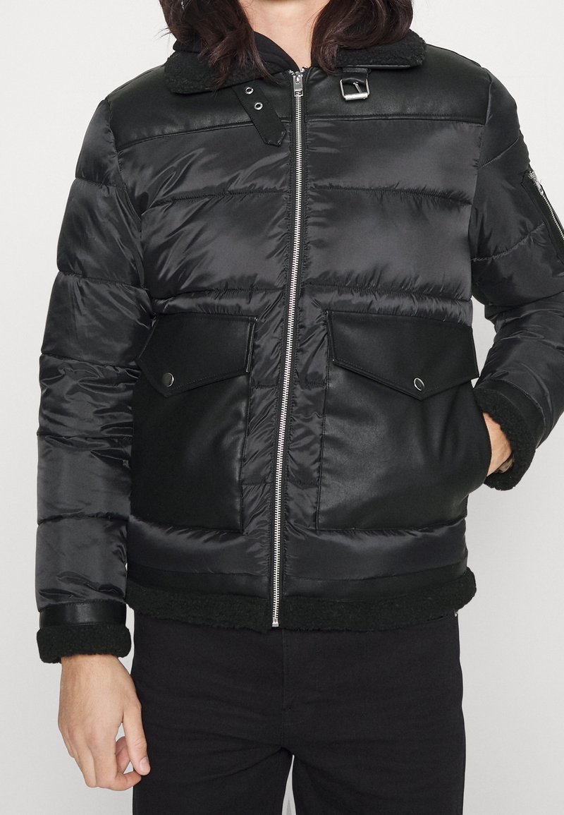 Personne portant une veste matelassée noire avec fermeture éclair à l'avant, deux grandes poches à rabat, col avec boucle, et une main dans la poche.