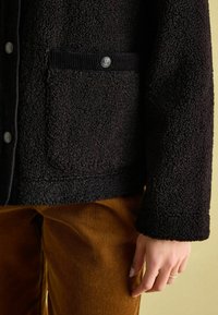 Czarna, textured fleece kurtka z kordurową kieszenią, srebrnym detalem guzikowym i luźnym krojem. Zestawiona ze złoto-brązowymi spodniami kordurowymi.