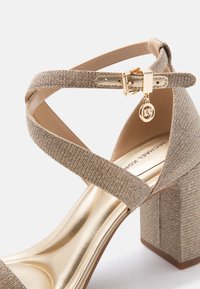 Σανδάλι με χοντρό τακούνι σε υφή χρυσού, με σταυρωτούς ιμάντες, αγκράφα στον αστράγαλο και με τη διακοσμητική πινελιά λογότυπου Michael Kors σε λευκό φόντο.