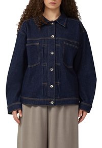 Femme portant une veste en jean bleu foncé avec des boutons sur le devant et deux poches poitrine, associée à un pantalon ample gris clair.