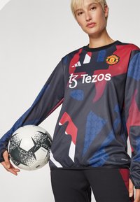 Manchester United-drakt i mørkt stoff med abstrakte røde og blå mønstre. Inkluderer logo og Tezos-merke, kombinert med en svart-hvit ball.