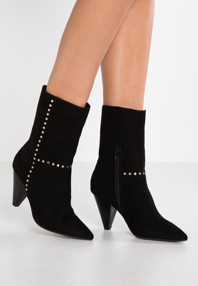 Bottes en daim noir mi-mollet avec bouts pointus, talons coniques, fermetures éclair latérales et détails en clous argentés portées sur jambes nues.