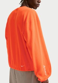 Άνδρας που φοράει φωτεινή πορτοκαλί φλις μπλούζα Polartec με μακρυά μανίκια και μικρό λευκό λογότυπο Nike κοντά στη μέση, εμφανιζόμενος από πίσω.