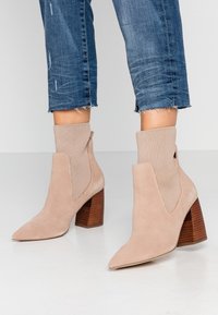 Beżowe zamszowe botki za kostkę z ostrym noskiem, dzianinową cholewką przypominającą skarpetkę oraz brązowym, piętą w kształcie słupka. W designie znajdują się boczne wycięcia.