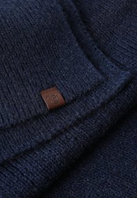 Tumši zils adīts džemperis ar reljefu rakstu, salocīts ar brūnu ādas birku, kuras apakšmalā iešūts lāča logotips.