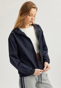 Marinblå lättviktsvindjacka med dragkedja framtill, huva och elastiska mudd. Matcher med ljusgrå sweatpants med sidosträck.