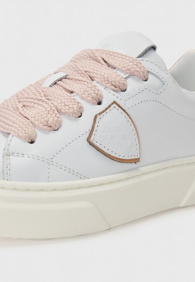 Hvitt lær sneakers med flettede rosa skolisser, brun logo-patch, teksturert gummisåle og minimalistisk design.