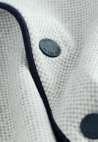 Grauer Textilstoff mit karierter Struktur, in Marineblau eingefasst, versehen mit einem runden, dunklen Knopf, der mit "Ted Baker" geprägt ist.