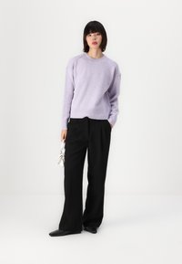 Pull lilas avec des manches raglan, texture douce et ourlet en côtes, associé à un pantalon noir à jambes larges et un petit sac à main blanc.