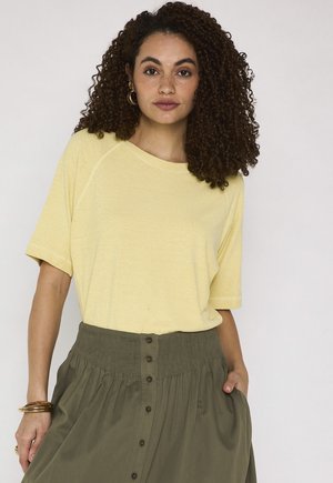 Femme aux cheveux bouclés portant un haut jaune à manches courtes et une jupe taille haute vert olive boutonnée, mains dans les poches.