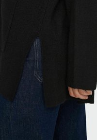 Svart stickad tröja med sidoklyftor, texturerad tyg och långa ärmar, tillsammans med mörkblå jeans med kontrasterande sömmar.