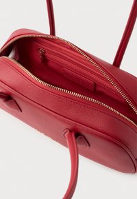 Červená kožená kabelka s texturovaným povrchem, vybavená zipovým uzávěrem a vnitřní kapsou. Zlaté zipové příslušenství a detaily šití.