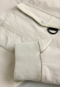 Tessuto del giubbotto beige con una finitura testurizzata, dotato di chiusura a bottoni, tasca a patta e polsini a coste di un colore simile chiaro.