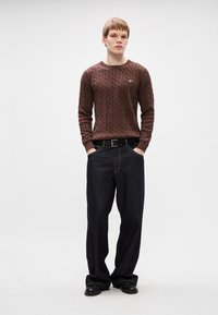 Σκούρο καφέ πουλόβερ σε πλέξη καμπάνας με ριγέ λαιμόκοψη, συνδυασμένο με σκούρους τζιν παντελόνες χαλαρής γραμμής και μαύρη ζώνη, φορεμένο με μαύρα παπούτσια.