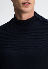 Man in een donkerblauwe geribde trui met hoge kraag en drie zwarte knopen op de linkerschouder.