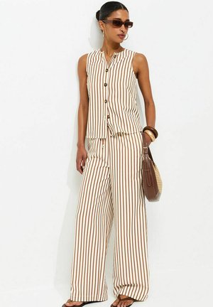 Vrouw die een beige mouwloze top met knopen draagt en een bijpassende wijde broek met verticale bruine strepen, houdt een bruine handtas vast en draagt een zonnebril.