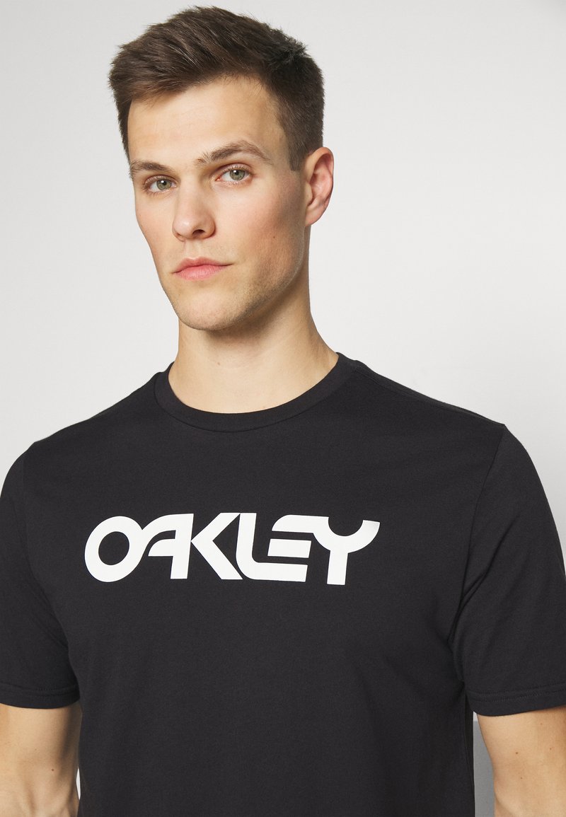 Camiseta de algodón negra con un prominente logo blanco de "OAKLEY" en el pecho. Mangas cortas y un diseño de cuello redondo estándar.