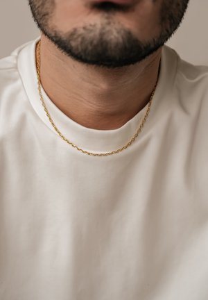 Goldene Halskette mit verknüpften ovalen Formen, getragen über einem schlichten weißen Rundhals-T-Shirt. Nahaufnahme, die Struktur und Glanz zeigt.