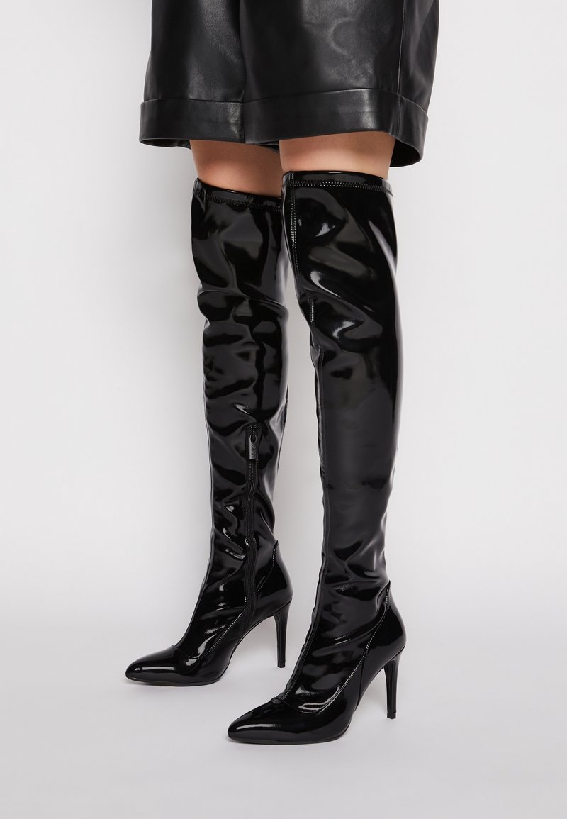 Botas de charol negro de caña alta con punteras afiladas y tacones de aguja. Cuentan con un cierre lateral y una textura lisa y brillante.