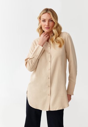 Camicia beige a maniche lunghe con taglio dritto, spacchi laterali e texture liscia, caratterizzata da un colletto a punta e sette bottoni rotondi.