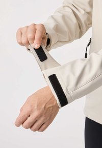 Main ajustant une bande velcro noire sur une manche de veste beige sur un fond blanc uni.