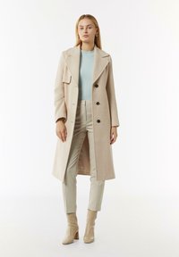 Beige wollblend jas met grote revers en vier zwarte knopen, gedragen over een mintgroene top en aangemeten crèmekleurige broek, gecombineerd met lichtbeige laarzen.